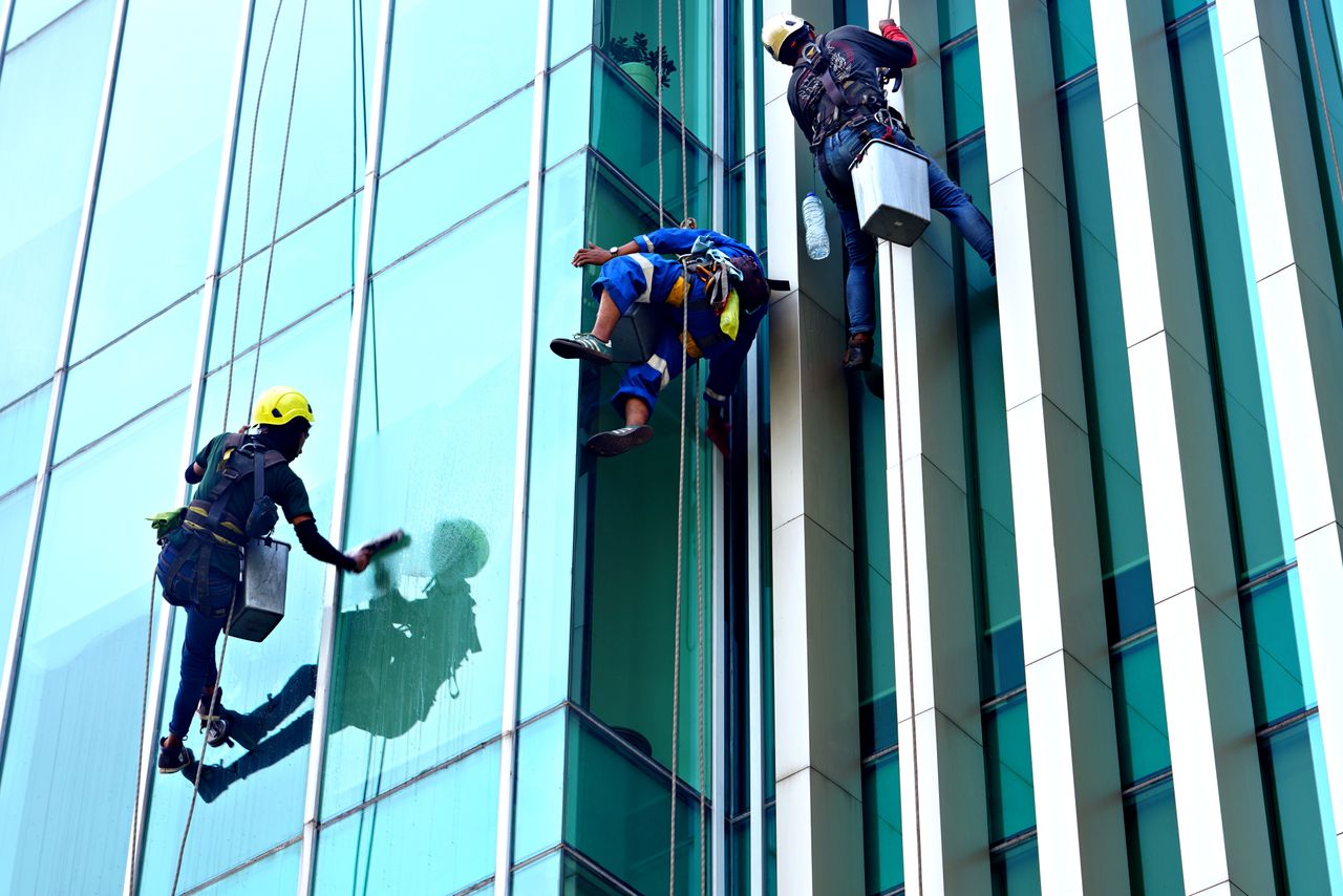 about-02 Three window cleaners suspended on ropes clean a modern glass building facade during the day.