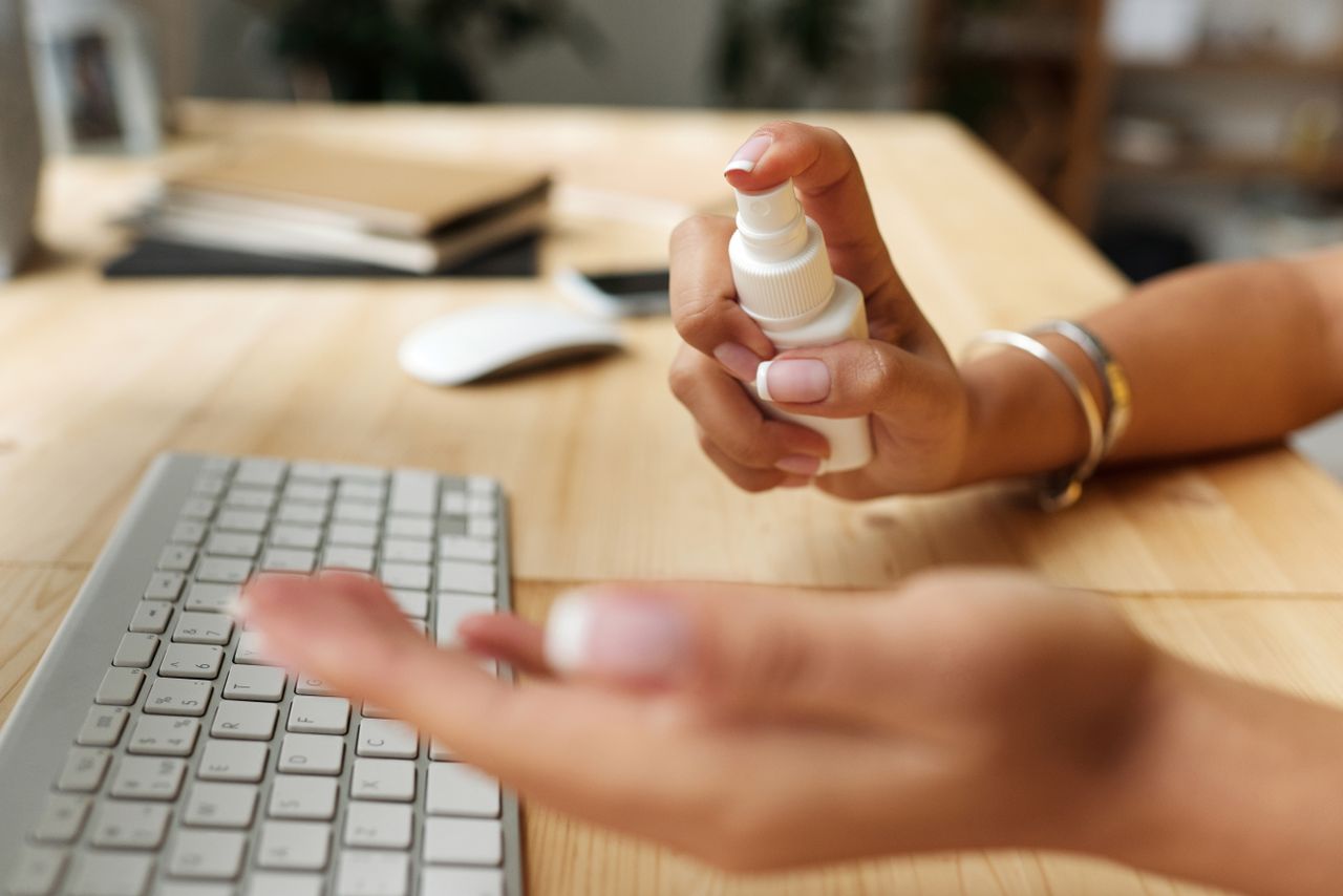 services-01 Person sanitizing hands at a desk with spray bottle, keyboard, and computer mouse.